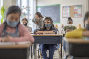 Group of elementary school classmates wearing protective face masks in the classroom while desks are socially distanced due to new COVID-19 regulations.